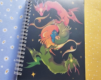 Może przedstawiać: Czarny notes w spirali z kolorową ilustracją dwóch splecionych ze sobą smoków. Smoki są zielone, pomarańczowe i czerwone z małym niebieskim stworzeniem w środku. Tło jest czarne z złotymi gwiazdami.