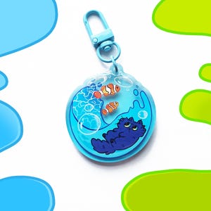 May include: A blue acrylic keychain featuring a cartoon cat and two clownfish in a water-filled bubble. The keychain has a light blue clasp. The cat is dark blue, and the fish are orange and white. The background is white with blue and green accents.