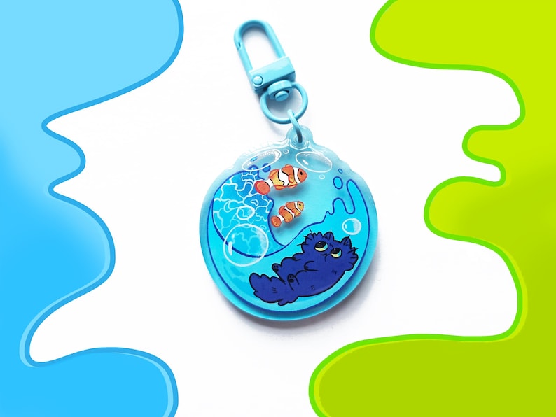 May include: A blue acrylic keychain featuring a cartoon cat and two clownfish in a water-filled bubble. The keychain has a light blue clasp. The cat is dark blue, and the fish are orange and white. The background is white with blue and green accents.