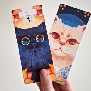 May include: Two colorful bookmarks featuring illustrations of cats. The bookmark on the left features a black cat with blue eyes and a yellow background. The bookmark on the right features a white cat with red eyes and a blue and white background.
