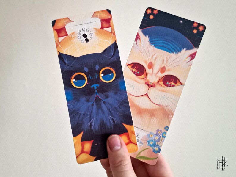 May include: Two colorful bookmarks featuring illustrations of cats. The bookmark on the left features a black cat with blue eyes and a yellow background. The bookmark on the right features a white cat with red eyes and a blue and white background.