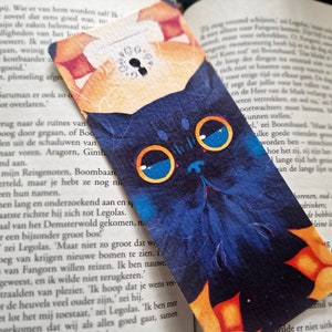 May include: A blue and orange bookmark with a black cat illustration. The cat has large blue eyes and a white nose. The bookmark has a keyhole design in the upper right corner.