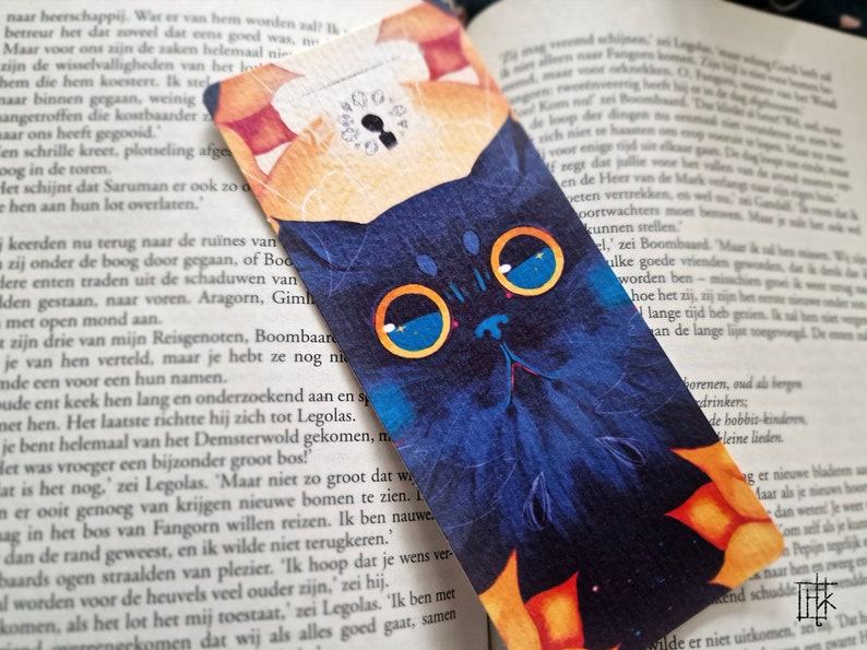 May include: A blue and orange bookmark with a black cat illustration. The cat has large blue eyes and a white nose. The bookmark has a keyhole design in the upper right corner.