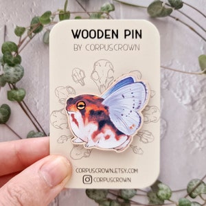 Op de afbeelding: Houten pin met een grillige illustratie van een kikker met vlinder vleugels. De kikker is geschilderd in tinten van oranje, rood, wit en zwart. De vleugels zijn lichtblauw met witte stippen. De pin is op een kaart met de tekst "WOODEN PIN BY CORPUSCROWN" en het website adres "CORPUSCROWN.ETSY.COM".