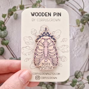 May include: Wooden pin with a pink and white anatomical ribcage design and the text "Death Positive". The pin is surrounded by a wreath of leaves and is on a white card with the text "Wooden Pin by Corpuscrown" and the website address "corpuscrown.etsy.com".