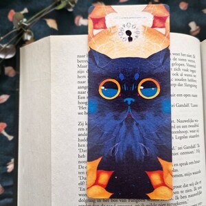 May include: A blue cat bookmark with large, round, yellow-rimmed glasses. The cat's eyes are blue with white stars. The bookmark has a yellow and orange background with a black keyhole in the upper right corner.