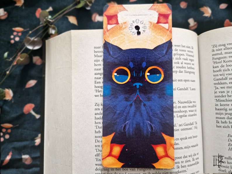 May include: A blue cat bookmark with large, round, yellow-rimmed glasses. The cat's eyes are blue with white stars. The bookmark has a yellow and orange background with a black keyhole in the upper right corner.