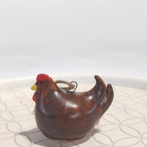 May include: A brown ceramic hen figurine with a red comb and yellow beak. The hen has a loop of twine for hanging. The figurine is a decorative item, possibly an ornament or kitchen decor.