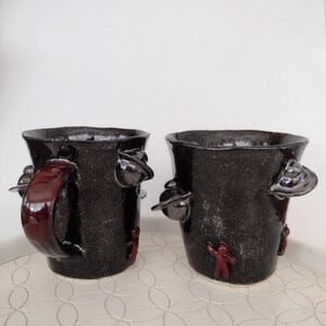 May include: Two handcrafted ceramic mugs with a dark, textured finish. Each mug features a unique design with red accents, including a handle on one and small human figures. The mugs have a rustic, artistic aesthetic.