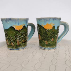 May include: Two ceramic mugs featuring a hand-painted landscape design. The mugs display a bright yellow sun rising over dark green pine trees, set against a light blue sky. Each mug has a sturdy handle and a slightly irregular, textured rim.