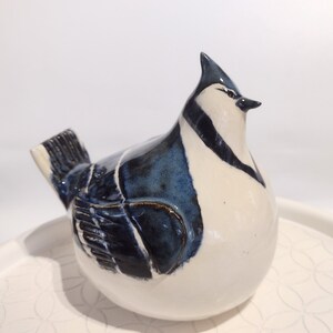 May include: A ceramic blue jay figurine with white and blue accents. The bird is sitting on a white surface with a decorative pattern.