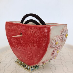 May include: A red and white ceramic dish with a floral design. The dish has a small hole in the side and is holding two black hair ties.
