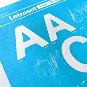 May include: A close-up of a blue sheet of paper with the text "Letraset Instant Lettering" printed in white. The letters "AA" are printed in white on the blue sheet.