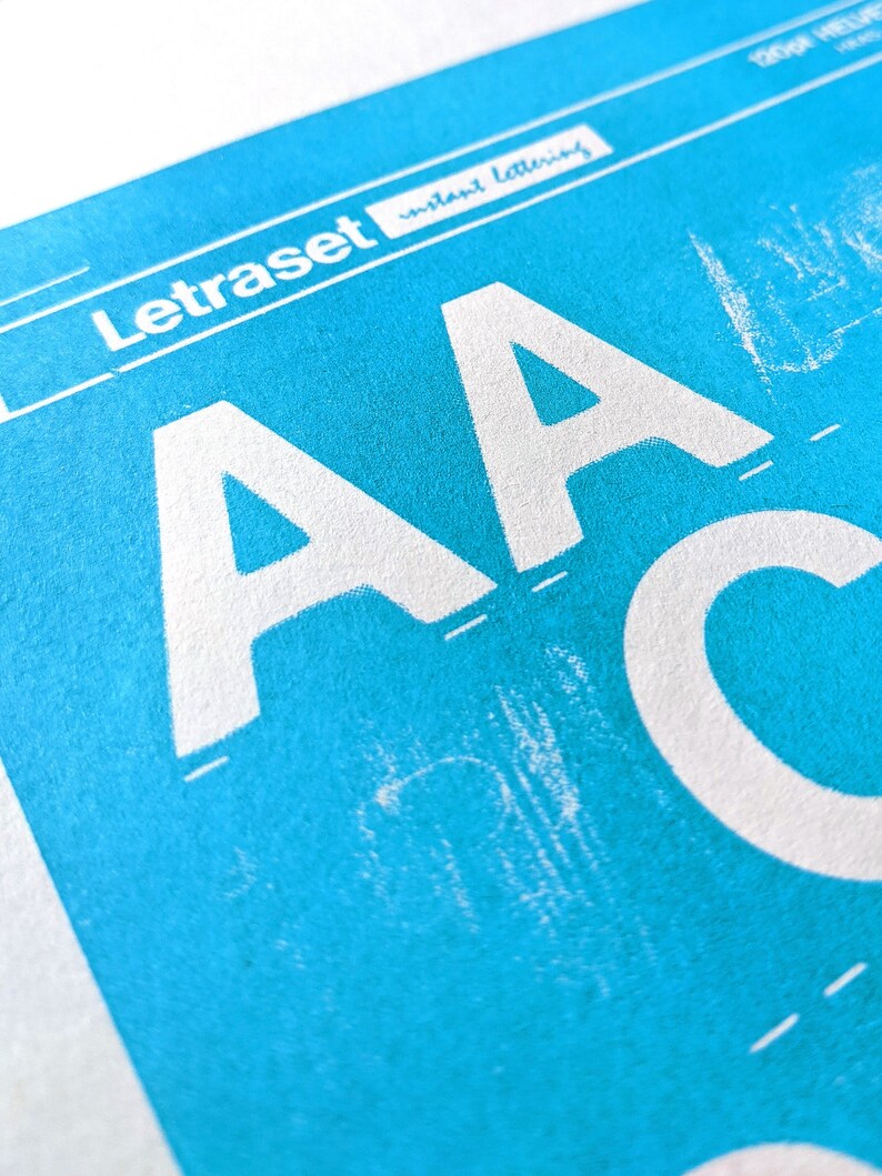 May include: A close-up of a blue sheet of paper with the text "Letraset Instant Lettering" printed in white. The letters "AA" are printed in white on the blue sheet.