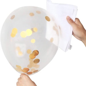 May include: A clear balloon filled with gold confetti. The balloon is being held by a hand, and a white cloth is being used to rub the balloon to help the confetti stick to the sides.