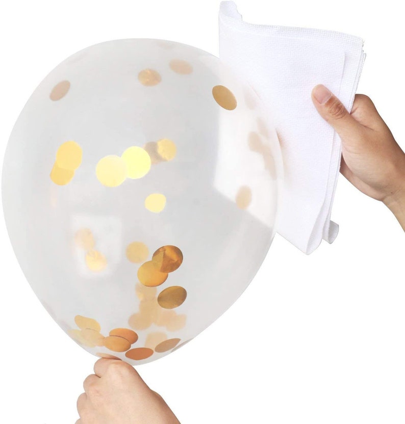 May include: A clear balloon filled with gold confetti. The balloon is being held by a hand, and a white cloth is being used to rub the balloon to help the confetti stick to the sides.