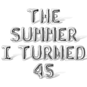 May include: Silver foil balloon letters spell out "THE SUMMER I TURNED 45" on a white background. The balloons are inflated and casting shadows, creating a celebratory and festive atmosphere.