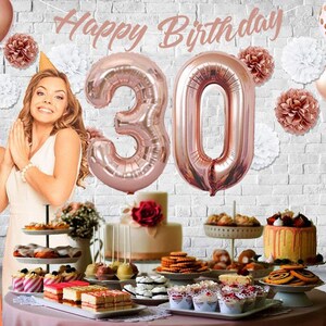 May include: A birthday party table setting with a rose gold theme. The table is decorated with rose gold balloons, including two large number balloons that spell out "30". There are also rose gold paper pom-poms and a banner that says "Happy Birthday". The table is set with a variety of desserts, including cupcakes, cake pops, and cookies.