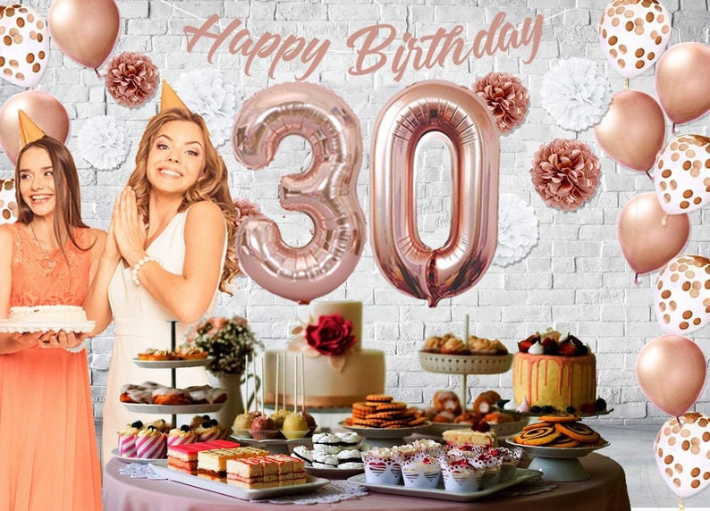 May include: A birthday party table setting with a rose gold theme. The table is decorated with rose gold balloons, including two large number balloons that spell out "30". There are also rose gold paper pom-poms and a banner that says "Happy Birthday". The table is set with a variety of desserts, including cupcakes, cake pops, and cookies.