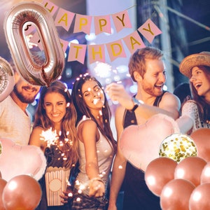 May include: A group of people celebrating a 60th birthday with rose gold balloons, confetti balloons, and a pink banner that says "Happy Birthday".