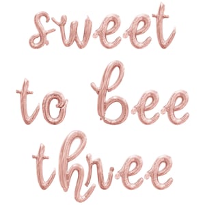 May include: Rose gold foil balloons spelling out "sweet to bee three" against a white background. The balloons are in a cursive font, perfect for a third birthday celebration. The balloons are inflated and ready to be displayed.