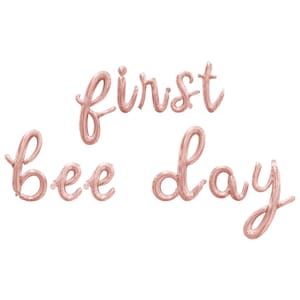 May include: Rose gold foil balloons spelling out "first bee day" in a cursive font. The balloons are arranged in three lines, with "first" on top, "bee" in the middle, and "day" at the bottom. The balloons are against a white background.
