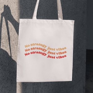 May include: White tote bag with the text "No strategy just vibes" in orange and red lettering. The bag is held by a hand against a textured gray wall.