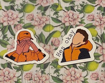 以下が含まれることがあります： 人物のイラストが描かれたステッカー2枚。最初のステッカーは、オレンジ色のジャケットとベースボールキャップを着用し、手で目を覆っている人物を描いています。2枚目のステッカーは、オレンジ色のジャケットと黒いフード付きのスウェットシャツを着用し、頭の上に「Hes okay」という文字が書かれた人物を描いています。