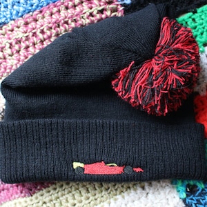 Puede incluir: Gorro negro con un pompón rojo y negro y una imagen bordada de un coche de carreras amarillo.