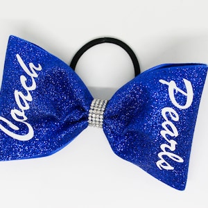 Op de afbeelding: Een blauwe glitter cheer bow met een wit elastiek en strass steentjes. De strik heeft de woorden "Coach" en "Pearls" in wit op elke zijde geschreven.