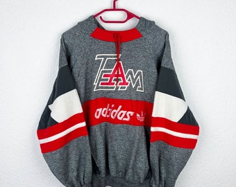 May include: Gray hooded sweatshirt with red and white color blocking. The sweatshirt has the word "TEAM" in red and white letters and the word "adidas" in red letters with the adidas logo.