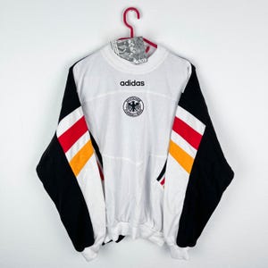 Op de afbeelding: Wit Adidas-sweatshirt met zwarte mouwen en rood-geel-witte strepen. Het sweatshirt heeft het Adidas-logo en een Duits nationaal team embleem. De mouwen zijn zwart met rood, geel en witte strepen. Het sweatshirt hangt aan een rode hanger.