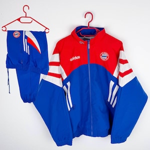 Könnte beinhalten: Ein Vintage Adidas Trainingsanzug in Blau, Rot und Weiß. Die Jacke hat einen durchgehenden Reißverschluss, das Adidas-Logo und ein Team-Emblem. Die passende Hose ist blau mit weißen Streifen und einem Team-Logo. Beide Teile hängen an roten Kleiderbügeln.