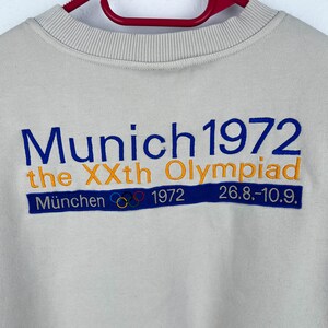 May include: A beige sweatshirt with blue and orange embroidered text that reads "Munich 1972 the XXth Olympiad" and "M&uuml;nchen 1972 26.8.-10.9.". The text is surrounded by five interlocking rings in blue, yellow, black, green, and red.