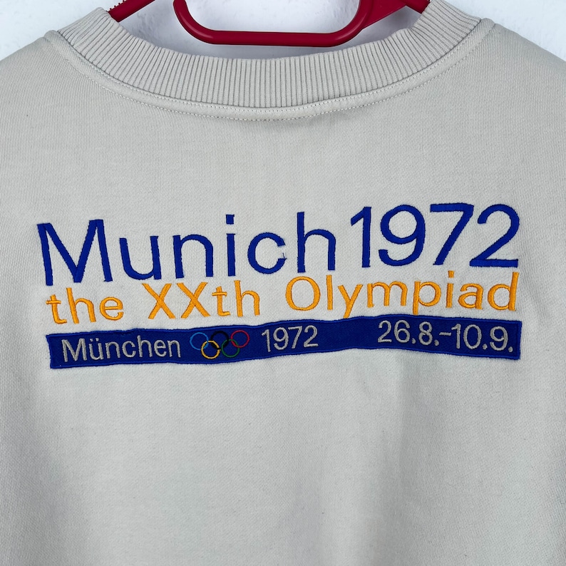 May include: A beige sweatshirt with blue and orange embroidered text that reads "Munich 1972 the XXth Olympiad" and "M&uuml;nchen 1972 26.8.-10.9.". The text is surrounded by five interlocking rings in blue, yellow, black, green, and red.