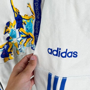 May include: White Adidas sweatshirt with blue stripes and the Adidas logo on the front. The sweatshirt has a graphic of basketball players on the left side with the text "Olympische Spiele Mun" on the side.