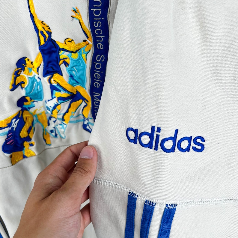 May include: White Adidas sweatshirt with blue stripes and the Adidas logo on the front. The sweatshirt has a graphic of basketball players on the left side with the text "Olympische Spiele Mun" on the side.