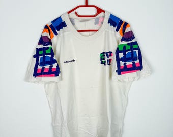 May include: White t-shirt with colorful abstract designs on the sleeves. The shirt has the Adidas logo on the left side and the text "E.T. 1982" on the right side.