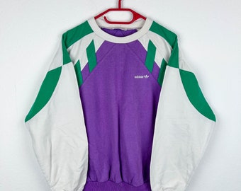 May include: A purple sweatshirt with white and green color blocking. The sweatshirt has a white crew neck and the Adidas logo on the front.