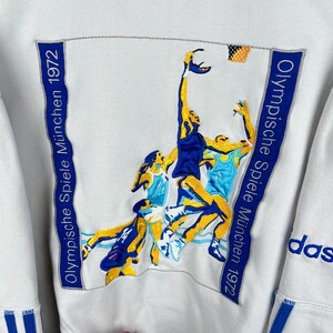 May include: White sweatshirt with a blue and yellow embroidered image of basketball players. The text "Olympische Spiele M&uuml;nchen 1972" is embroidered on the right side of the image. The word "adidas" is embroidered on the right sleeve.