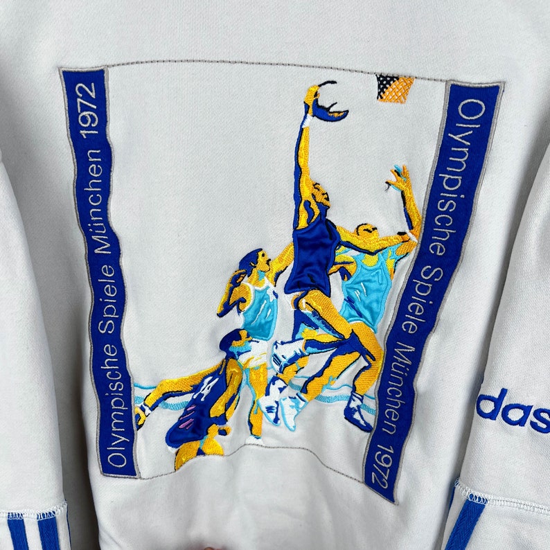May include: White sweatshirt with a blue and yellow embroidered image of basketball players. The text "Olympische Spiele M&uuml;nchen 1972" is embroidered on the right side of the image. The word "adidas" is embroidered on the right sleeve.