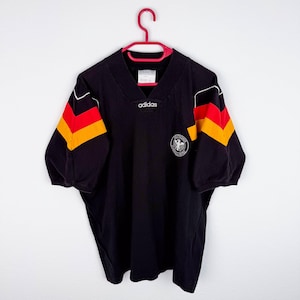 May include: A black short-sleeved jersey with a V-neck collar featuring the Adidas logo at the centre. The sleeves display horizontal red and yellow stripes. A circular emblem with a white eagle and the text Deutscher Fussball-Bund is on the chest.