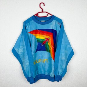 Könnte beinhalten: Ein blaues Sweatshirt mit einem Regenbogen und einer Skifahrergrafik. Das Sweatshirt hat den Text "Ridge Lift" auf der Vorderseite gestickt.