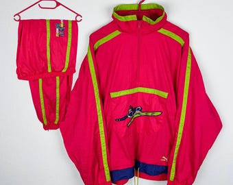 May include: A pink and green vintage Puma tracksuit with a skier graphic on the jacket. The jacket has a half zip closure and a drawstring hood. The trousers have an elastic waistband and a drawstring.