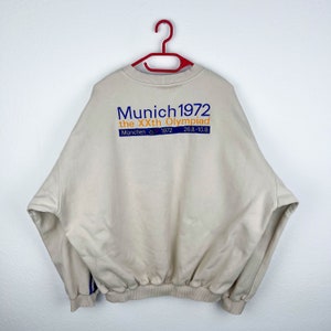 May include: A beige sweatshirt with a blue and orange graphic print that reads "Munich 1972 the XXth Olympiad Munich 1972 26.8-10.9."