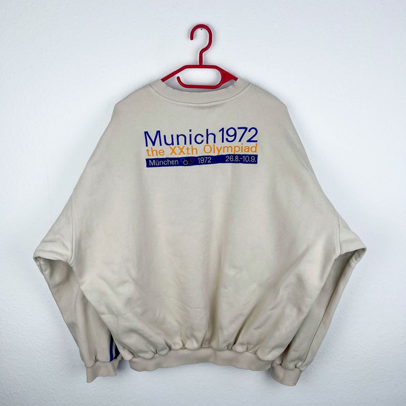 May include: A beige sweatshirt with a blue and orange graphic print that reads "Munich 1972 the XXth Olympiad Munich 1972 26.8-10.9."