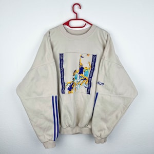 May include: A beige sweatshirt with blue and white stripes on the sleeves. The front of the sweatshirt features a graphic of athletes competing in the 1972 Munich Olympics with the text "Olympische Spiele M&uuml;nchen 1972" and the Adidas logo.