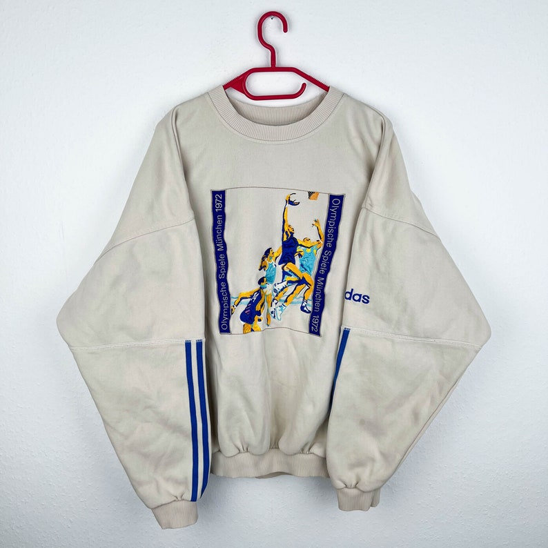May include: A beige sweatshirt with blue and white stripes on the sleeves. The front of the sweatshirt features a graphic of athletes competing in the 1972 Munich Olympics with the text "Olympische Spiele M&uuml;nchen 1972" and the Adidas logo.