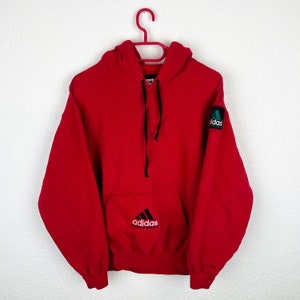 Puede incluir: Sudadera roja con capucha y cordones negros, con un logotipo Adidas Equipment en blanco y negro en el bolsillo delantero. La sudadera también tiene un parche con el logotipo de Adidas en verde y negro en la manga derecha.