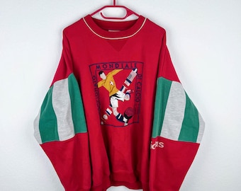 Könnte beinhalten: Roter Adidas-Sweatshirt mit grün-grauen Farbblockärmeln. Der Sweatshirt zeigt eine gestickte Grafik eines Fußballspielers mit dem Text "MONDIALE DI CALCIO" um die Grafik herum.
